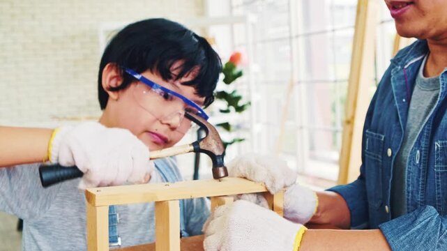 Happy Asian father and son work as a woodworker and carpenter. Father teaching his son to hammer nails on a wooden plank carefully together. carpentry working at a home workshop studio.