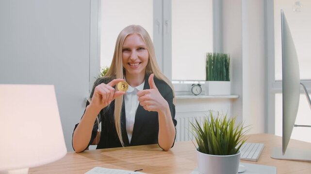 A smiling young blonde woman in a dark blazer holds a gold bitcoin and gives a thumbs up while seated at a desk in a bright modern office.