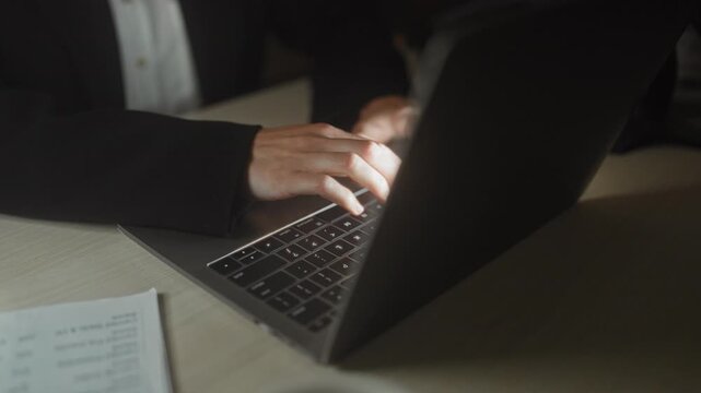 Woman typing on laptop in office at night, hands on keyboard with nearby papers and blazer cuff visible on desk; late night focus.