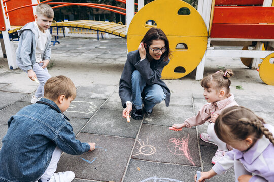 Positive smiling teacher and young children drawing on playground with chalk