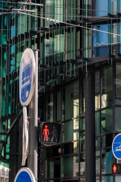 Red pedestrian signals and traffic lights punctuate an urban street intersection, standing against the layered glass facade of a contemporary building