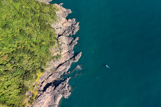Lush forest covers the rugged rocky coastline of Son Tra Peninsula in Danang as a small boat sails across the calm turquoise ocean water on a clear sunny day