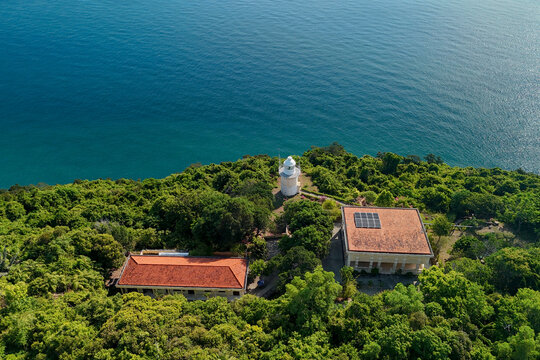 Green forest covers the rocky cliffs surrounding a white lighthouse and buildings overlooking the calm blue sea on Son Tra Peninsula in Danang