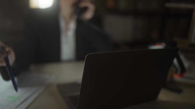 Woman holding telephone receiver to ear at desk in building, reaching for laptop and pen with face lit by lamp; focused professionalism.