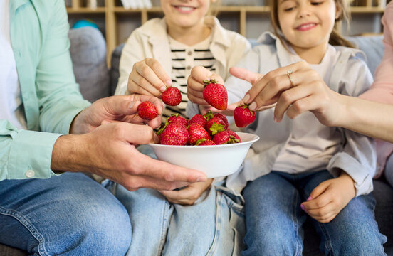 Family children eating strawberries at home together. Parents and kids share fresh fruit on the couch, promoting nutrition and habits. Healthy family snack and bonding concept at home.