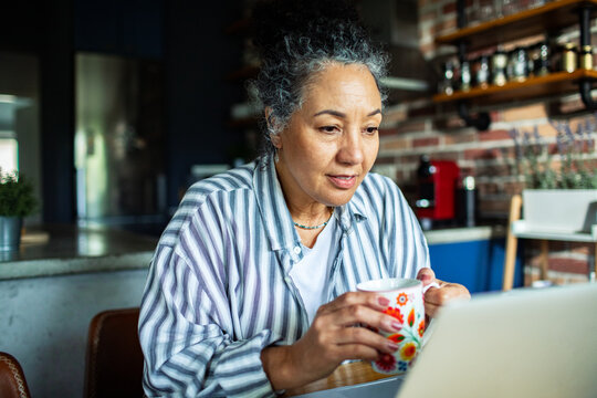Mature woman working on laptop with coffee at home kitchen