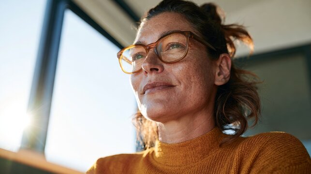 Woman with glasses looking out a window with her hair in a bun, wearing an orange top and sunglasses.