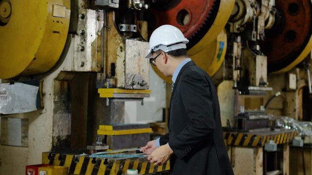Confident Asian male engineer in suit and hard hat inspecting metal parts at manufacturing plant. Industrial factory manager smiling with crossed arms standing by heavy machinery equipment production.