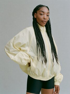 Black woman with long braids wearing a yellow windbreaker jacket and black shorts poses confidently against a neutral gray background, showcasing modern fashion style