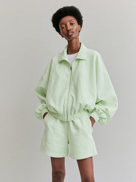 African woman wearing a sage green lightweight zip-up jacket and matching shorts poses confidently against a neutral background, showcasing a modern casual fashion style