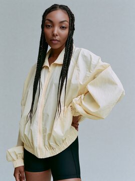 Black woman with long braids wearing a light yellow oversized jacket and black shorts poses confidently against a neutral background, showcasing modern fashion style