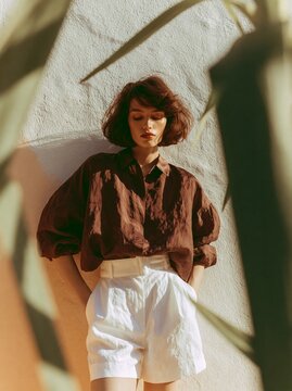 Woman in rich chocolate brown satin oversized shirt and white shorts poses against a textured wall, framed by green foliage, showcasing a stylish summer look