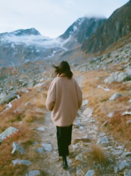 Woman in oversized camel jacket walks along a rocky path in mountainous terrain with grass and snow patches, showcasing a natural outdoor landscape