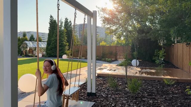 Teenage girl with long red hair rides a wooden swing and turns toward the camera while wearing wireless headphones in slow motion. Concept of confidence, authenticity and positive self expression.