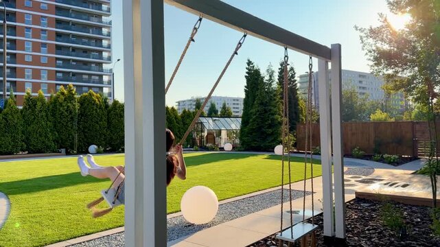 Teenage girl with red hair smiles while swinging outdoors with wireless headphones around her neck as her hair moves in wind in slow motion. Concept of freedom, femininity and joyful summer lifestyle