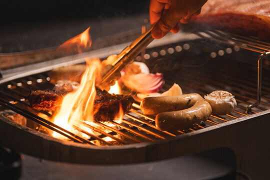 Flame grill barbecue sausage meat tong hand onion garlic fire, close cooking scene with food over metal grate, smoky heat, outdoor kitchen, warm light, rustic dinner preparation