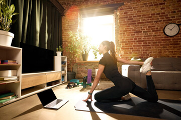 Obraz premium Woman stretching quadriceps on mat during online home workout. Concept of post-workout recovery, muscle flexibility, injury prevention, mobility improvement and virtual fitness coaching sessions.