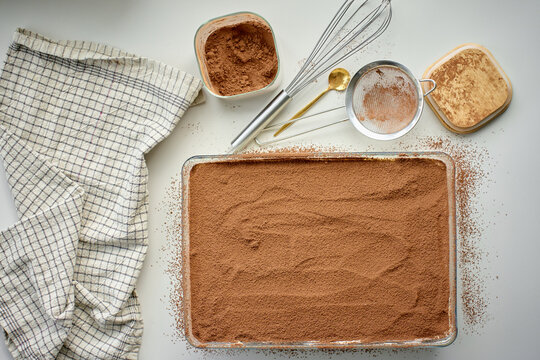 Homemade tiramisu dessert dusted with cocoa in dish, with sieve and kitchen towel nearby