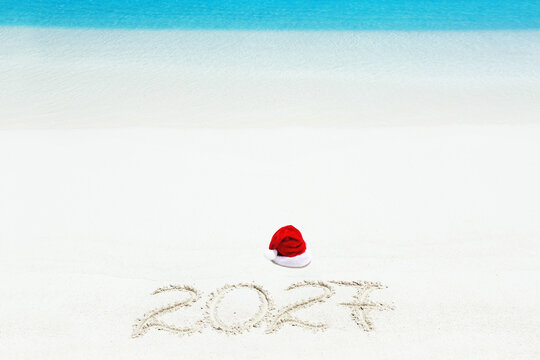 Santa Claus hat and 2027 inscription written on sandy beach, New Year card. Christmas celebration on seashore