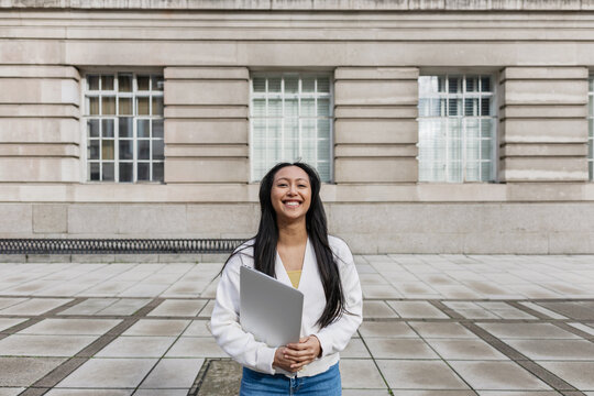 Woman Student smiling while holding laptop on campus