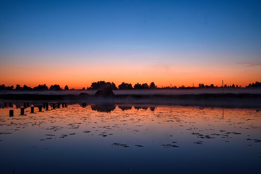 Misty lake at sunrise with old wooden pier posts, calm reflections, silhouetted trees, and a colorful dawn sky.