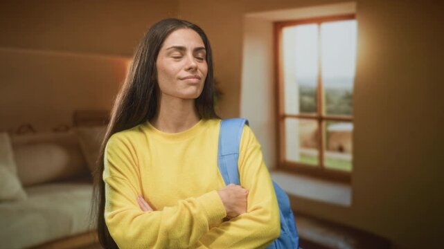 Woman with backpack and yellow sweater finger to temple and arms folded inside a building room by a window and sofa; thoughtful uncertainty.
