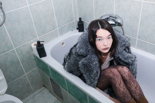 Young Person Sits in Bathtub Wearing a Coat and Looking Thoughtful