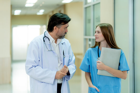 Experienced doctor and young nurse discussing patient care in a modern hospital corridor highlighting teamwork clinical communication and quality healthcare delivery.

