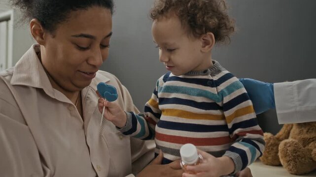 Biracial mother and young child visiting pediatrician in clinic, child holding medicine bottle and lollipop. Footage is perfect for pediatric healthcare and family wellness campaigns