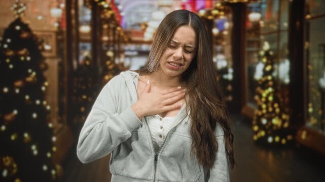Woman clutches chest for heartattack, wincing and holding hand to sternum in a decorated shopping building with holiday lights; distress concern.