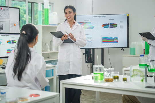 Laboratory scientist presentation report screen chart woman researcher in white coat explaining experiment data beside glassware with green liquid in modern classroom