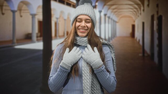 Woman in knit hat, scarf and gloves places gloved hands on chest while smiling in historic building corridor; cozy joy.