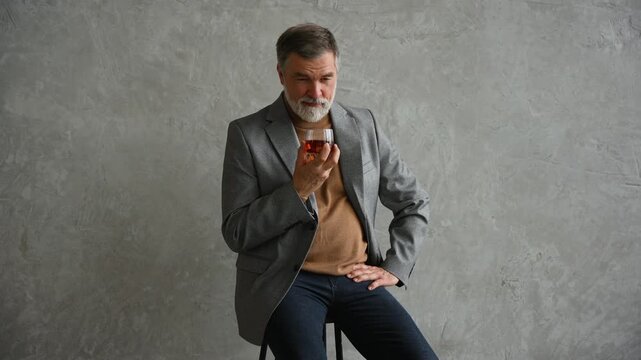 Handsome and successful businessman in stylish suit holding glass whiskey while sitting on the stool
