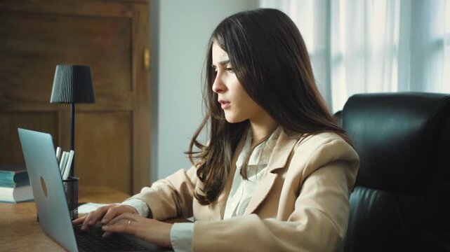 Young Hispanic businesswoman sneezing while working at her office desk, showing exhaustion and illness as she continues typing in a modern corporate workplace.
