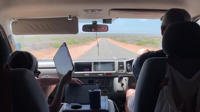 Inside a vehicle traveling on a long road, a driver focuses on the path while a passenger uses a tablet for entertainment.