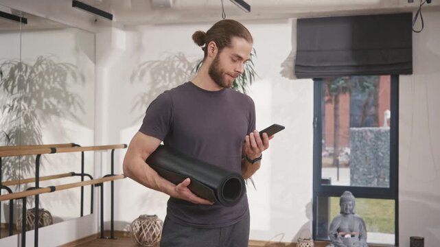 Yoga instructor standing at yoga studio. Sportive man holding yoga mat. male in sportswear holding smartphone and chatting. Yoga coach preparing for yoga session. Man practicing spiritual meditation.