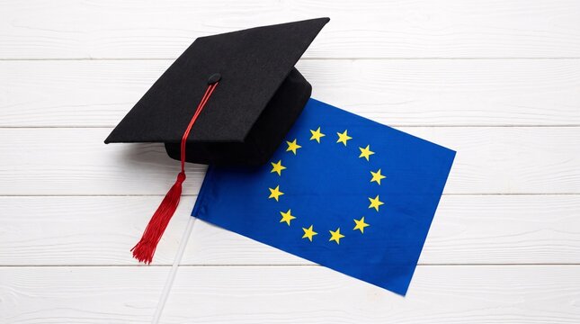 Black graduation cap with a red tassel resting beside a European Union flag on a textured white wooden surface symbolizing academic success