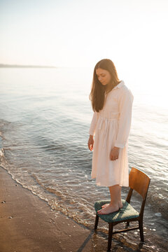 Beautiful young woman wearing white linen dress standing on chair in sea water at coastline. Loneliness.