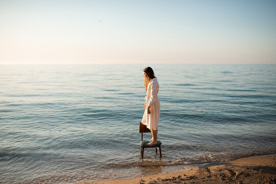 Lonely woman standing on chair in sea water wearing white linen dress at coast line. Loneliness.