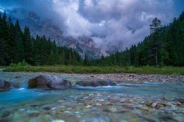 A beautiful mountain river flows through a dense coniferous forest under a dramatic, cloudy sky. Perfect for travel, nature, and environmental conservation themes. © marcodenaro
