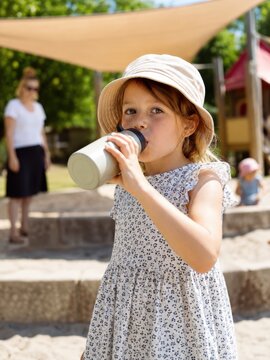 Kleines M&auml;dchen mit Sonnenhut trinkt bei hei&szlig;em Sommerwetter aus einer Trinkflasche. Schattensegel und leichte Kleidung bieten Schutz vor Hitze und starker Sonne.