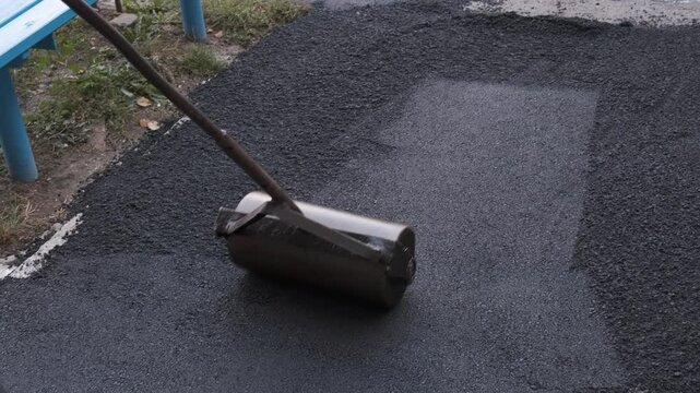 Close-up of a worker using a heavy manual metal roller tool to flatten and compact the edge of freshly laid hot asphalt next to a concrete curb.