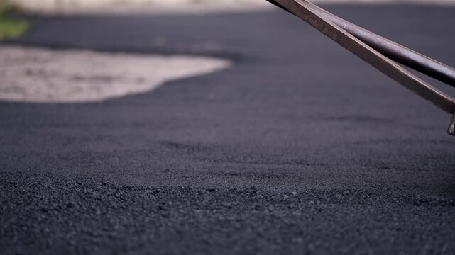 A closeup shot of a hand-operated metal roller compacting and smoothing a freshly laid layer of hot black asphalt on a pavement walkway.