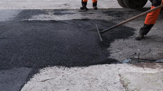 A close-up shot of a road worker's legs and rake leveling hot, granular black asphalt during pavement repair and maintenance works.