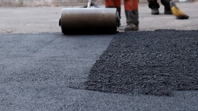 A low-angle, close-up shot of a heavy steel hand roller tool compacting freshly laid hot black asphalt with a worker standing in the background.