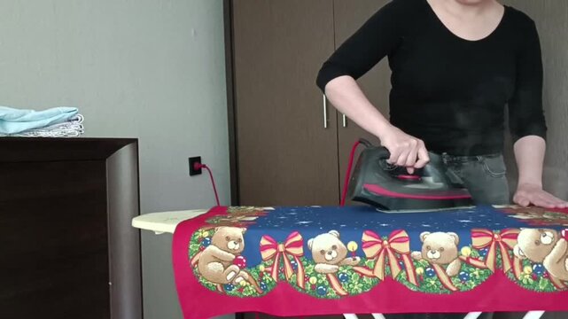 Person ironing a Christmas-themed teddy bear fabric with star patterns. Hands pressing iron across colorful cloth on board, bedroom background with dresser.