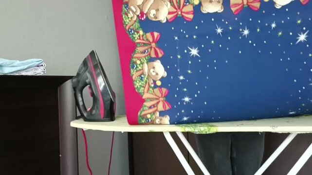 Hands folding a festive Christmas tablecloth with teddy bears and stars on an ironing board. Neatly folded laundry on dresser, wardrobe in background.