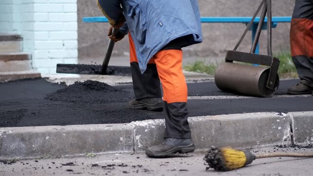 Workers in orange protective trousers leveling new black asphalt pavement using a special wide rake on a city street.