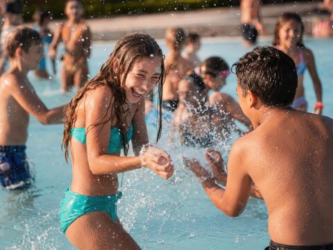 Zwei Jugendliche liefern sich lachend eine ausgelassene Wasserschlacht im gut besuchten deutschen Freibad. Fliegende Tropfen und Bewegung vermitteln pure Sommerfreude.