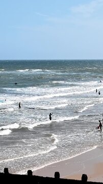 Vertical panning shot from a high view over Cromer beach, following people enjoying the cool waves on a bright sunny day, with the sandy shoreline, sparkling sea and North Norfolk coast below.